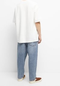 Persona vistiendo una camiseta blanca holgada, jeans claros y anchos, y zapatos marrones de deslizamiento, de pie contra un fondo blanco liso.
