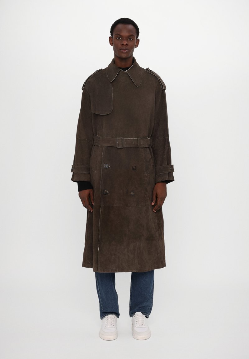 Brauner Wildleder-Trenchcoat mit doppelt geknöpftem Design, großem Kragen, Schulterepauletten und Gürtel. Getragen über einem schwarzen Hemd und blauen Jeans.