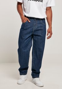Denim jeans in dark blue with a loose fit, featuring a button fly, five pockets, and white stitching. Paired with white sneakers.