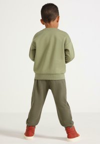 Felpa verde oliva con collo rotondo e polsini a costine, abbinata a pantaloni verde oliva larghi e testurizzati e stivaletti rossi alla caviglia.