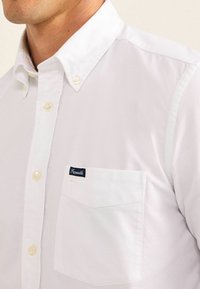 Camisa blanca de cuello, estilo abotonado con botones visibles, un solo bolsillo en el pecho y una pequeña etiqueta azul oscura cosida en el bolsillo.