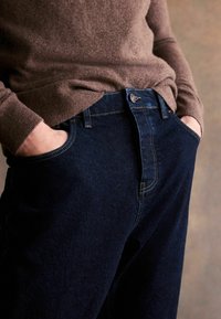 Jeans din denim albastru închis, cu o croială dreaptă, prezentând un buton și un fermoar vizibile, și două buzunare frontale, asociate cu un pulover maro.