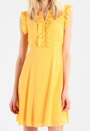 Mouwloze gele jurk met kleine witte stippen, gerimpelde kraag, knopenlijst en getailleerde taille, gedragen door een staande persoon.
