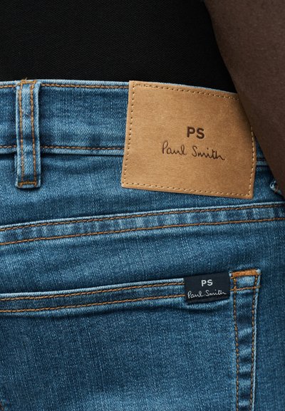 Κοντινό πλάνο μπλε τζιν με καφέ δερμάτινη ετικέτα Paul Smith στη μέση και μαύρη υφασμάτινη ετικέτα στην πίσω τσέπη.