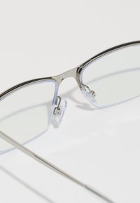 Óculos de metal com acabamento prateado, lentes retangulares, almofadas nasais transparentes e varas finas que se estendem da armação.