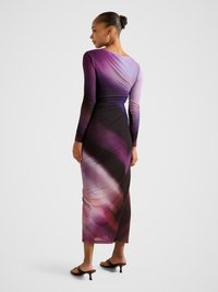 Moteris su surištais plaukais, dėvinti ilgomis rankovėmis prigludusią vijoliškai juodą gradientinę maxi suknelę ir dėvinčia rudus aukštakulnius sandalus, stovinti priešais vienspalvį foną.