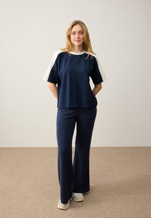 Vrouw staat met handen achter de rug, draagt marineblauwe wijde broekspijpen, marineblauwe korte mouw top met witte schouderpanelen en beige sneakers.