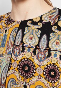 Gros plan sur le cou et l'épaule d'une femme portant un vêtement coloré à motif cachemire avec des dessins complexes en jaune, noir, bleu et rouge.