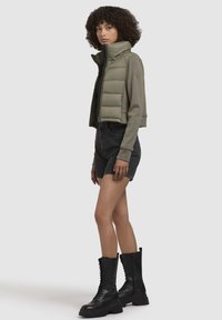 Jachetă croppată puffy, de culoare verde măslin, cu guler înalt, purtată cu pantaloni scurți negri din denim și cizmulițe negre cu șireturi până la genunchi. Materiale fine și texturate.