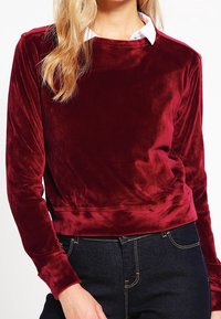Suéter de terciopelo burdeos con mangas largas y escote redondeado, superpuesto a una camisa blanca con cuello. Textura suave y lisa.