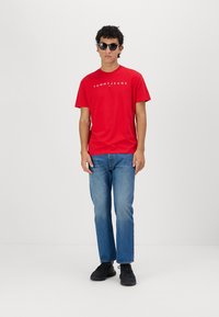 T-shirt en coton rouge avec le logo "TOMMY JEANS", associé à un jean en denim bleu et des baskets noires. Le modèle porte des lunettes de soleil noires.