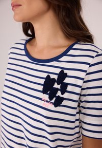 Gestreiftes Baumwoll-T-Shirt mit dunkelblauen und weißen Linien. Verziert mit strukturierten, dunkelblauen, gestickten Akzenten und pinkem Text mit der Aufschrift "GEBE LIEBE ZURÜCK."