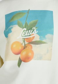 Fehér póló, amelyen színes narancsok zöld levelekkel láthatók, kék ég és felhő háttér előtt, a "Levi's" logóval.