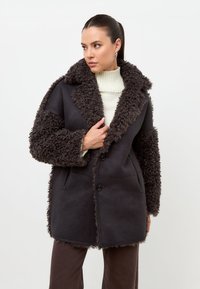 Kvinde iført mørkebrun lammeskindspels med tekstureret krave og manchetter over hvid rullekrave, kigger til siden, stående mod en hvid baggrund.