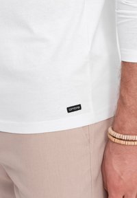 Weißes Langarm-T-Shirt aus Baumwolle, mit einem kleinen schwarzen "Ombre"-Label am unteren Saum, kombiniert mit beigefarbenen Hosen und Holzarmbändern.