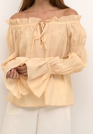 Kvinde iført en lysebeige off-shoulder bluse med flæsekant ved halsudskæringen, pufærmer og udvidede manchetter, kombineret med hvide bukser.