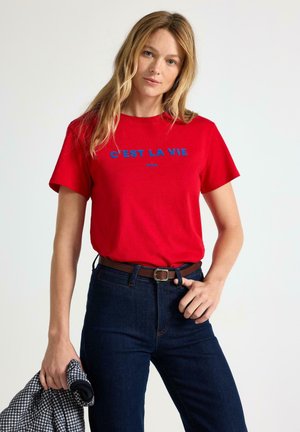 Kvinde iført en rød T-shirt med blå tekst "C'EST LA VIE PARIS,", mørke jeans og et brunt bælte, der holder en sort-hvid ternet skjorte.