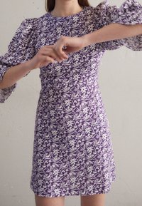 Robe violette avec un motif floral blanc, dotée de manches bouffantes et d'une silhouette ajustée, fabriquée en tissu léger.