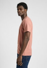 Różowa bawełniana koszulka z okrągłym dekoltem, krótkimi rękawami i dopasowanym krojem, w zestawie z ciemnoniebieskimi dżinsami. Widoczna marka na piersi.