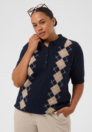 Femme aux cheveux foncés attachés porte des lunettes de soleil sur la tête, un polo bleu marine à motif losanges beige, et un pantalon clair, posant avec une main près du visage.