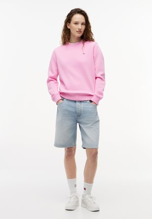 Mladá osoba stojaca s rukami v kapsách, oblečená v ružovej mikine, svetlých denimových šortkách, bielych ponožkách a bielych teniskách na bielom pozadí.