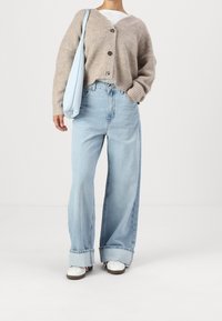 Béžový pletený cardigan s velkými knoflíky, světle modrá taška, oversize světle modré džínsy a bílé tenisky s červenými a hnědými akcenty.