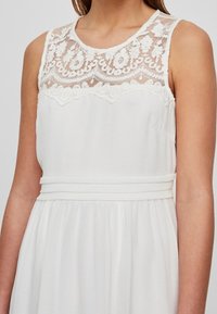 Robe blanche présentant un corsage en dentelle avec des motifs floraux, un col rond et une taille cintrée. La jupe est fluide et texturée.