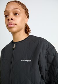 Crna prešivena jakna s patentnim zatvaračem, s okruglom ovratnikom i Carhartt logotipom na prsima. Glatka tekstura i minimalistički dizajn.