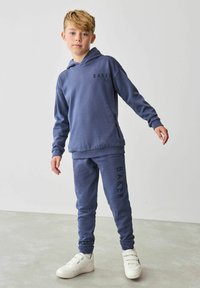 Modrá mikina s kapucňou a zodpovedajúce tepláky vyrobené z mäkkej tkaniny; na hrudi a na boku sa nachádza logo. Topánky sú biele a majú viacero popruhov.