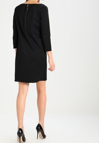 Robe noire mi-longue avec des manches trois-quarts, dotée d'une fermeture éclair au dos et d'un col rond. Texture lisse et design ajusté.