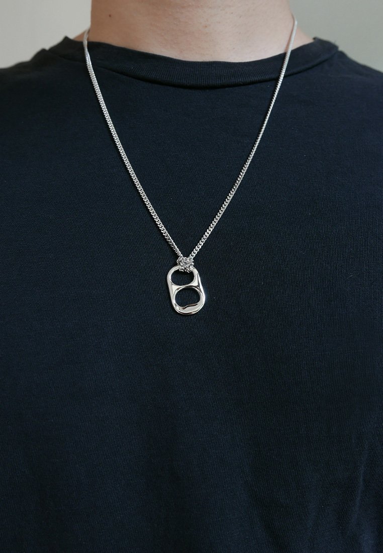 Collana d'argento con una catena texturizzata, che presenta un pendente unico e astratto che assomiglia a un anello aperto. Indossata sopra una t-shirt nera.