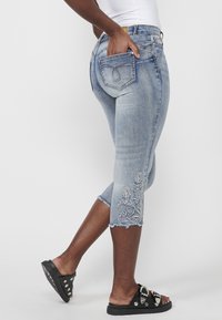 Personne portant un jean court bleu clair brodé et des sandales noires slip-on avec des boucles argentées décoratives et des clous, debout de profil.