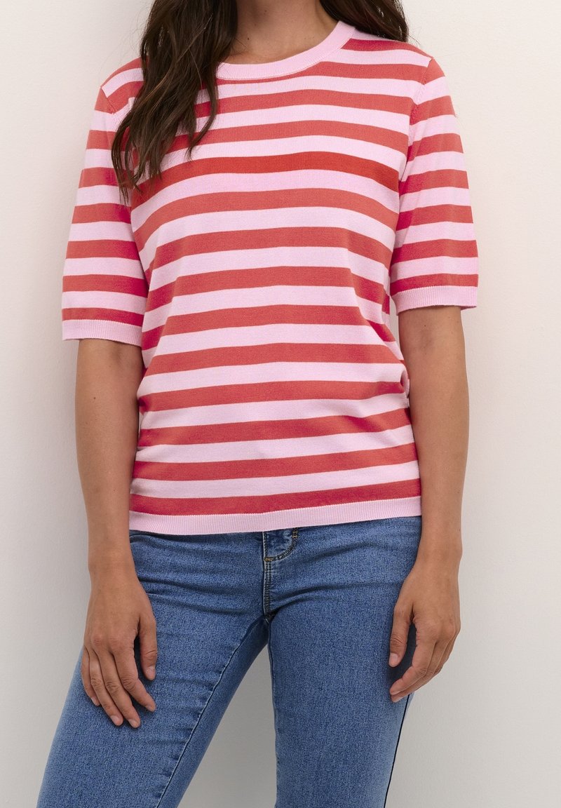 Mujer que lleva una camiseta de rayas horizontales en rosa y rojo con mangas cortas y jeans azules, de pie contra una pared blanca lisa.