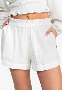 Shorts blancs texturés avec une taille élastique froncée et des poches latérales. Présente un ourlet légèrement retourné et un subtil détail en métal de la marque.