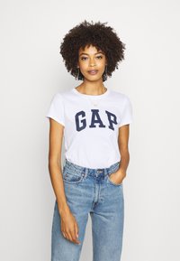 White cotton t-shirt with "GAP" in navy bold letters, round neckline, short sleeves, paired with high-waisted light blue denim jeans.