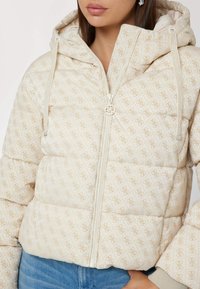 Frau trägt eine beige gesteppte Pufferjacke mit Kapuze, gemustert mit einem hellbraunen Monogramm, kombiniert mit blauen Jeans.
