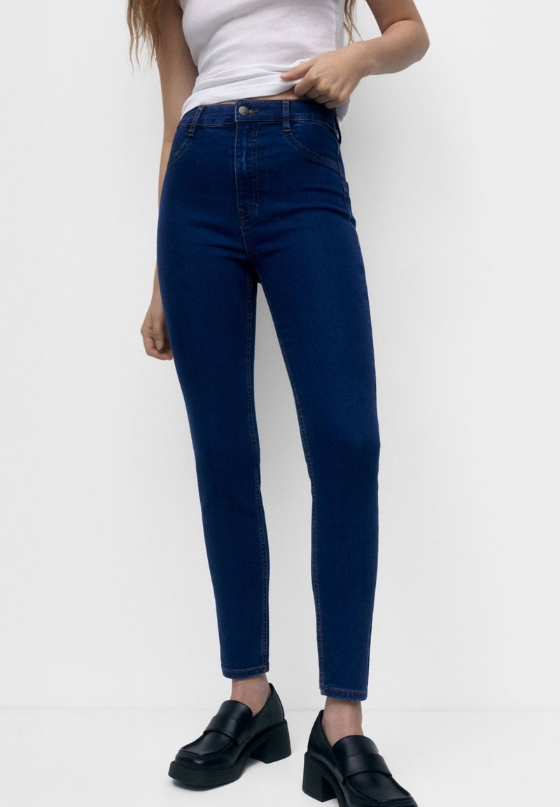 Jean skinny bleu taille haute en denim, avec fermeture éclair et bouton, cinq poches, et une texture lisse, légèrement extensible.