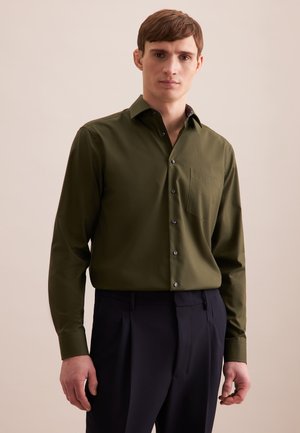 Chemise boutonnee vert olive à manches longues, avec une poche poitrine et des boutons noirs. Portée avec un pantalon foncé, sur un fond neutre.