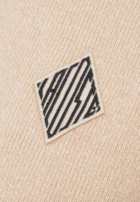 Bēšs audums ar teksturētu virsmu, uz kura ir melns izšūts ģeometriska dizaina logotips uz krēmkrāsas uzlīmes.