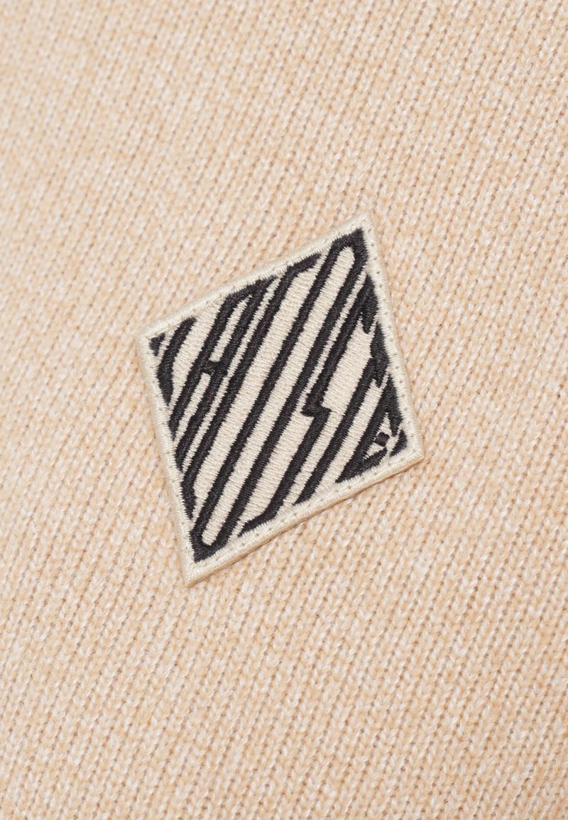 Beige stof met een gestructureerd oppervlak, voorzien van een zwart geborduurd logo in een geometrisch ontwerp op een crèmekleurig patch.