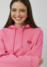 Sweat à capuche rose avec cordons et logo brodé « daily ». Texture douce, coupe décontractée, manches larges et poignets côtelés.
