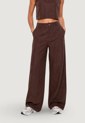 Persona che indossa pantaloni marrone scuro a vita alta e gamba larga, top corto senza maniche e scarpe da ginnastica bianche con le mani in tasca.