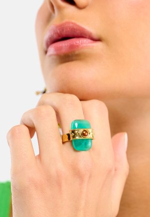 Bague en or ornée d'une grande pierre verte émeraude, accentuée par une petite pierre brune et des détails ornés sur l'anneau. Finition lisse.