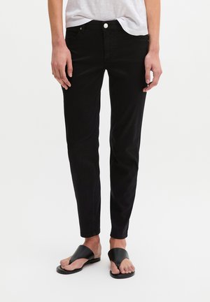 Personne portant un pantalon noir slim, une chemise blanche et des sandales noires à bout ouvert, debout sur un fond blanc uni.