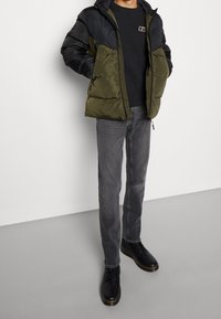 Pufjakke med olivengrønne og sorte paneler, lynlås lukning, parret med grå slim-fit jeans og sorte snørebåndsstøvler, på en grå baggrund.