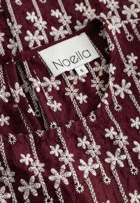 Tumepunane kangast pluus, millel on peen valge lillemotiiviga tikand, ümardatud kaelus ning silt kirjadega "Noella" ja suurusega "S". Kõikjal põhjalik õmblustöö.