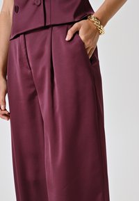 Pantaloni in satin bordeaux con una vestibilità rilassata, dotati di tasche laterali e pieghe sottili. Un bracciale dorato intrecciato adorna il polso.