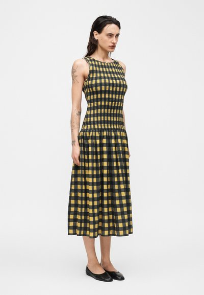 Γυναίκα που στέκεται με ένα μίντι φόρεμα χωρίς μανίκια, κίτρινο και μαύρο καρό, με εφαρμοστό πάνω μέρος και πιέτες στη φούστα, φοράει μαύρα μπαλαρίνια.