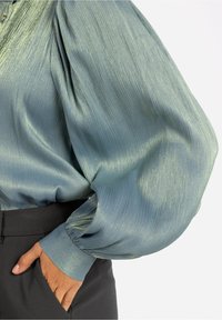 Blouse bleu clair à manches longues plissées avec des manches volumineuses, resserrées au poignet, rentrée dans un pantalon noir taille haute avec la main dans la poche.