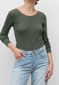 Langärmliges, olivgrünes Rippoberteil mit Rundhalsausschnitt, kombiniert mit hellblauen hochgeschnittenen Jeans. Das Material wirkt dehnbar und figurbetont.